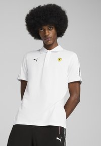 Hvid polo-shirt med krave, prydet med et gult Ferrari-logo og et sort Puma-logo. Korte ærmer med sorte accenter og mønstre.