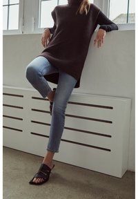 Suéter oscuro de cuello alto acanalado, jeans ajustados azul claro y sandalias negras con tiras. El fondo presenta listones horizontales blancos.