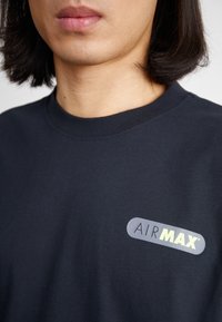 Navyblaues Baumwoll-T-Shirt mit Rundhalsausschnitt und einem hervorgehobenen grauen AIRMAX-Logo mit gelben Akzenten im linken Brustbereich.