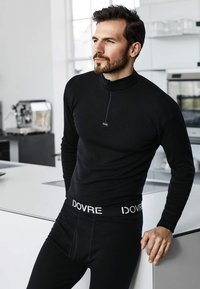 Sort termisk langærmet bluse med lynlås og logo, parret med sorte underdele med en hvid mærkebånd i taljen, glat tekstur.