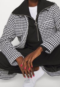 Giacca a motivo houndstooth bianco e nero con colletto nero e zip. Indossata sopra una maglia nera e un paio di pantaloni neri, abbinata a delle sneakers bianche.