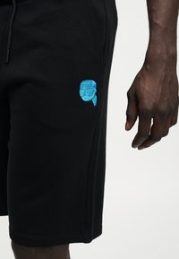 Μαύρο σορτς με μικρό μπλε κεντημένο λογότυπο ενός ατόμου που φορά γυαλιά ηλίου και μαντίλα, και ένα σκουρόχρωμο χέρι που ακουμπάει δίπλα τους.