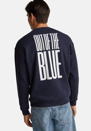 Man wearing navy sweatshirt with large white text "OUT OF THE BLUE" on the back, paired with light blue jeans.