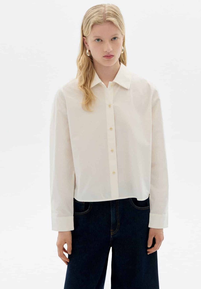 White button-up shirt with a collar, long sleeves, and a cropped design. Features yellow buttons and a smooth fabric texture.