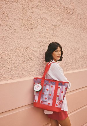 Õlgadeni ulatuvate tumedate juustega naine, kes kannab valget särki ja punast seelikut, toetub roosa tekstuurse seina vastu ja kannab suurt läbipaistvat punaste täppidega kotti.