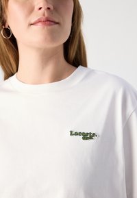 Hvit bomull T-skjorte med grønn brodert "Lacoste" logo og krokodille-design på venstre bryst. Korte ermer og rund hals.