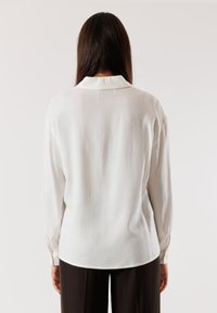 Camicia bianca a maniche lunghe con colletto, tessuto leggermente testurizzato, dotata di polsi bottone e una vestibilità rilassata, abbinata a pantaloni scuri.