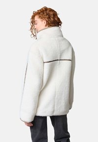 Vit fleecejacka med hög krage, oversized passform och subtila bruna accenter på axlarna, texturerat tyg och vida ärmar.