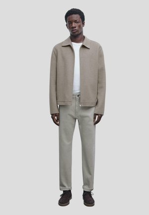 Beige wool jacket with a pointed collar, worn over a white T-shirt, paired with light gray trousers and dark brown shoes.