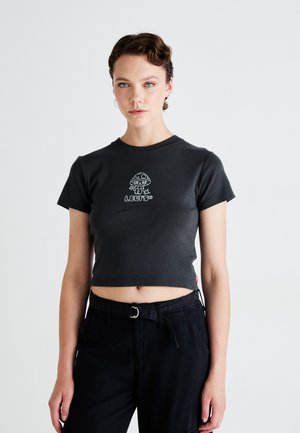 Jonge vrouw met kort, gekruld haar, gekleed in een zwart cropped t-shirt met een wit paddenstoelontwerp en hoge zwarte broek tegen een witte achtergrond.