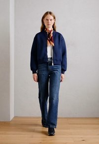Veste bomber courte bleu marine avec des poignets côtelés, portée sur un haut blanc et associée à un jean flare bleu taille haute et des chaussures noires.