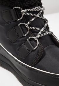 Black ankle boot made of textured fabric and leather, featuring round metal eyelets, white laces, and a contrasting rubber sole.