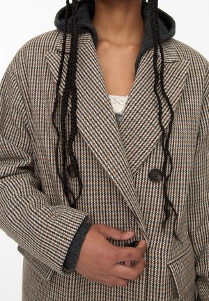 Osoba nosząca płaszcz w brązowo-beżowy wzór pepitki na ciemnej bluzie z kapturem, zapinająca przedni guzik płaszcza.