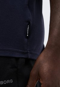 Κοντινή λήψη ενός σκοτεινόχρωμου χεριού δίπλα σε μπλε ναυτικό πουκάμισο Björn Borg και μαύρο παντελόνι με λογότυπο Björn Borg.