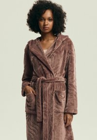 Różowy, pluszowy szlafrok z kapturem i długimi rękawami. Posiada dwie przednie kieszenie oraz pasek do zawiązania. Tkanina o fakturze z falistymi wzorami.