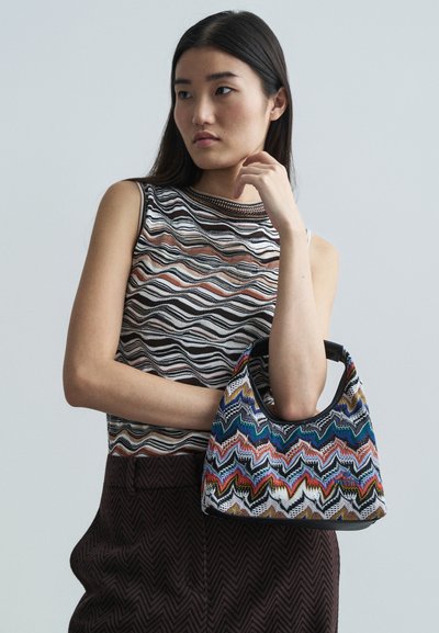 Textured, multicolored knit bag with zigzag patterns, featuring a black handle. Worn with a striped, sleeveless top and dark textured pants.