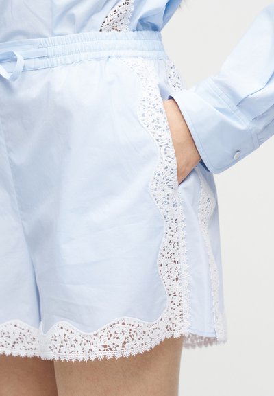 Ανοιχτό μπλε σορτς με λευκή δαντέλα κατά μήκος των πλευρών και του τελειώματος, με ελαστική μέση και ένα χέρι χαλαρά στην δεξιά τσέπη.