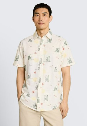 Man draagt een crèmekleurig kortärmelig knoopoverhemd met surfplank-, zon- en vogelprints, en beige broek, staand tegen een lichte achtergrond.