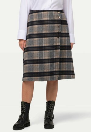 Kobieta w szarej spódnicy do kolan w kratę z czarnymi poziomymi paskami, białej bluzce i czarnych sznurowanych botkach stojąca.