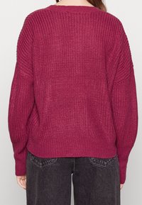 Maroon knit sweater with a loose fit, textured fabric, wide neckline, and ribbed hem, paired with dark denim pants. visible stitching details.