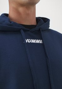 Haut à capuche marine avec un cordon de serrage et une poche kangourou à l'avant. Présente un logo blanc "HUMMER" sur la poitrine. Matière douce et texturée.