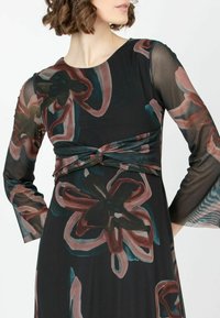 Blumengemustertes Kleid mit schwarzem, durchsichtigem Überzug, mit gerafftem Bund und langen Ärmeln. Die Farben umfassen Burgunderrot, Petrol und Creme.