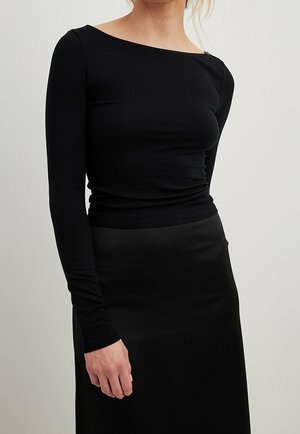 Mujer con un top negro ajustado de manga larga y escote alto barco, combinado con una falda negra, sobre un fondo neutro.