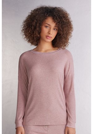 Vrouw met krullend haar die een licht mauve geribd shirt met lange mouwen en bijpassende broek draagt tegen een effen lichte achtergrond.
