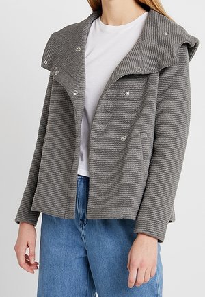 Femme portant une veste grise côtelée avec des boutons-pression argentés sur un t-shirt blanc et un jean bleu, vue du cou aux hanches.