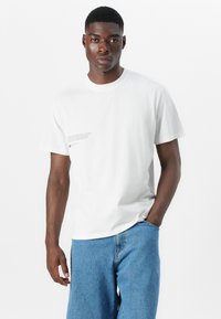 Weißes Baumwoll-T-Shirt mit rundem Ausschnitt, kurzen Ärmeln und bedruckten Textdetails auf der linken Seite. Kombiniert mit hellblauen Jeansshorts.