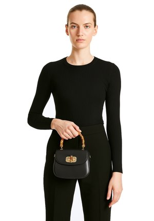 Sac à main en cuir noir avec un fermoir doré, forme structurée, anse courte avec des accents texturés. Le modèle porte un haut noir à manches longues à côtes.
