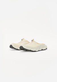 Klogg-liknande slip-on skor i krämfärgad nätmaterial med en texturerad yta, accentuerade av en svart gummisula och ett draghandtag.