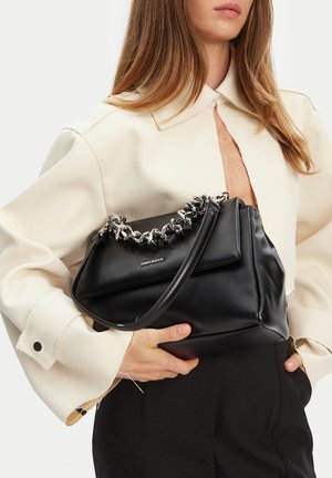 Donna che tiene una borsa nera in pelle con catena e tracolla argentate, indossa una giacca corta color crema e pantaloni neri.