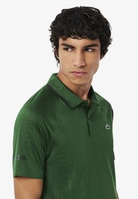 Polo verde in tessuto morbido con motivo geometrico testurizzato, rifiniture del colletto nere e logo Lacoste sul petto e sulla manica.