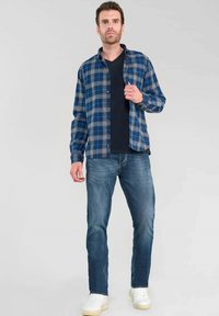 Camisa de franela a cuadros en azul combinada con una camiseta negra de escote en V, jeans de mezclilla y zapatillas blancas. La camisa cuenta con detalles de botones y mangas largas.
