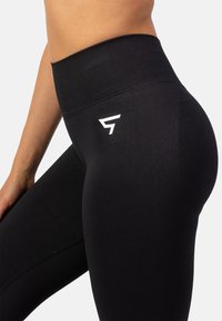 Čierne legíny s vysokým pásom a hladkou textúrou, s bielym logom na boku a zosilneným pásom pre podporu.