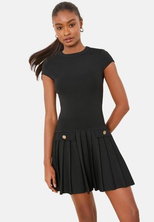 Kvinde med langt hår samlet i en hestehale iført en tætsiddende sort top med korte ærmer og et plisseret sort skørt med guldfarvede knapdetaljer.