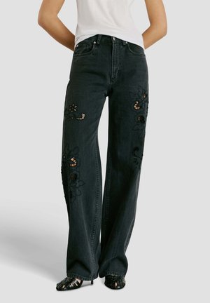 Persoon draagt een zwarte high-waist wijde broek met bloemenuitgesneden borduurwerk en zwarte sandalen met bandjes, tegen een effen achtergrond.