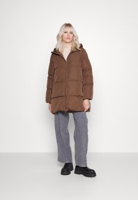 Palton pufos maro cu fermoar frontal și glugă. Material texturat cu un design matlasat. Asortat cu pantaloni largi gri și cizme negre.