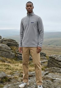 Mann steht auf felsigem Gelände, trägt eine graue Fleecejacke mit Viertel-Reißverschluss, khakifarbene Hose und hellfarbene Turnschuhe, im Hintergrund ein wolkiger Himmel.