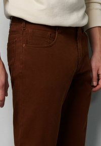 Braune Baumwollhosen mit glatter Textur, ausgestattet mit vorderen Taschen und einem klassischen Fünf-Taschen-Design. Der Bund hat Gürtelschlaufen.