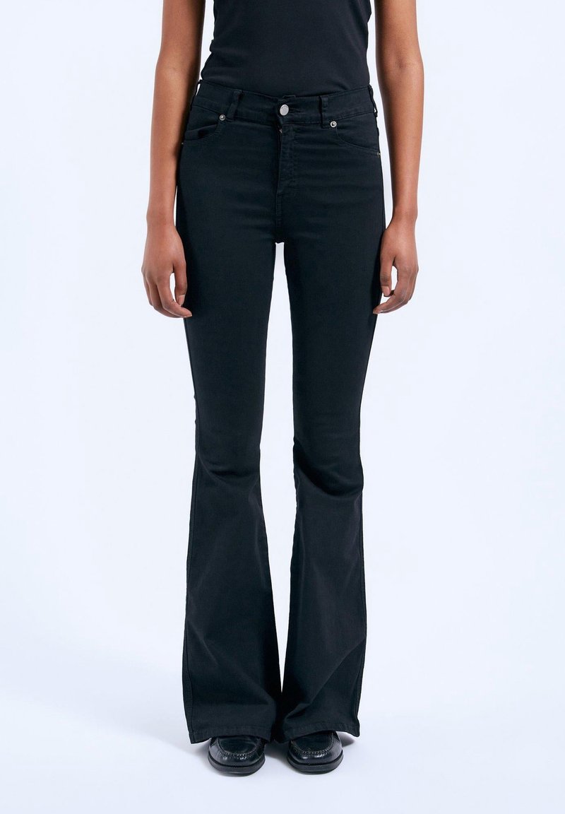 Personne portant un jean noir ajusté taille haute flare avec poches avant, fermeture à boutons, et chaussures noires sur un fond uni.