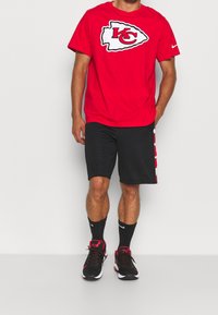 Camiseta roja de manga corta con el logotipo blanco y rojo "KC", combinada con pantalones cortos negros con acentos laterales blancos. Llevada con zapatillas negras y rojas.