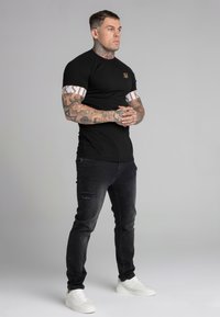 Melnas īsām piedurknēm t-krekls ar baltām un zelta akcentiem, apvienots ar nolietotām tumšām džinsām un baltām sporta kedām; redzamas tetovētas rokas.