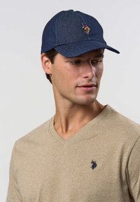 Dunkle Jeansmütze mit gebogenem Schirm und bestickten Logoakzenten. Getragen zu einem beige-farbenen V-Ausschnitt-Shirt. Nahaufnahme des Gesichtsprofils.