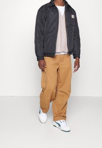 Černá prošívaná bunda s rozepínáním na zip a kapsou na boku, vrstvená přes béžovou pletenou vestu; doplněná béžovými cargo kalhotami a bílými teniskami.