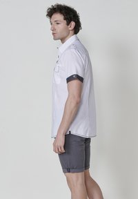 Camisa blanca de manga corta con cuello abotonado, acentos en azul marino en los puños, y pantalones cortos grises. Tejido suave con detalles de costura visibles.