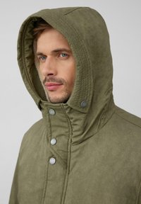 Man met een lichte baard die een olijfgroene gewatteerde jas met metalen drukknoopjes draagt, kijkt iets naar links tegen een effen achtergrond.