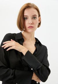 Camicia di seta nera con colletto, maniche lunghe e dettagli con bottoni. Texture liscia e vestibilità comoda. Braccio del modello parzialmente visibile.