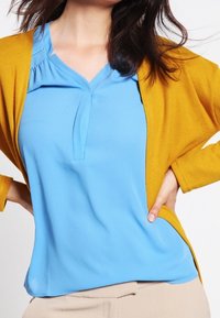 Femme portant un chemisier bleu à col en V avec un cardigan ouvert jaune moutarde et un pantalon beige, les mains posées sur les hanches.